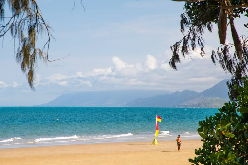 Une plage à l’eau d’un bleu limpide, avec un drapeau de sauveteur et des montagnes qui se dessinent à l’horizon.