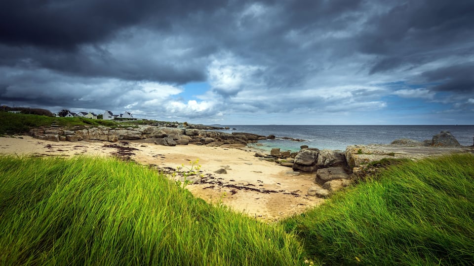 A coastal landscape with a rocky beach, grassy dunes, and a cloudy sky.