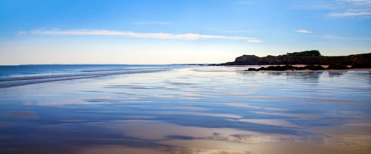 A wide, calm beach with gentle waves, a clear sky, and rocky cliffs in the background.