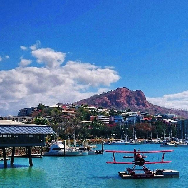 A coastal scene with a mountain in the background, a harbor with boats, and a seaplane on the water.