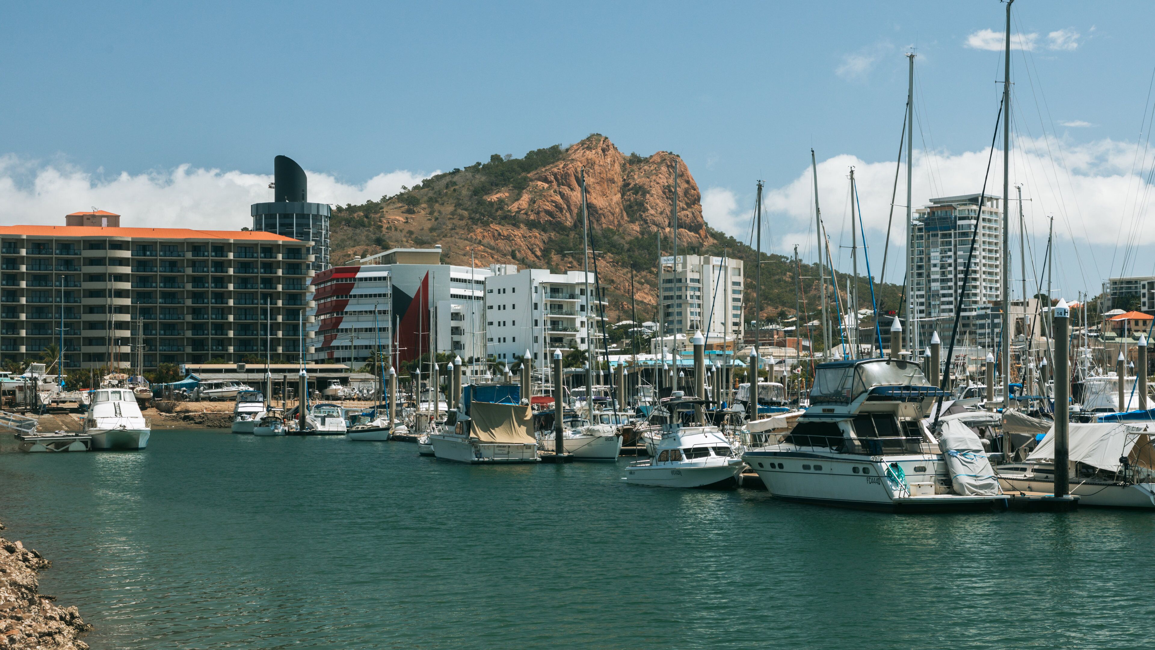 A marina with boats docked, a mountain in the background, and buildings along the shore.