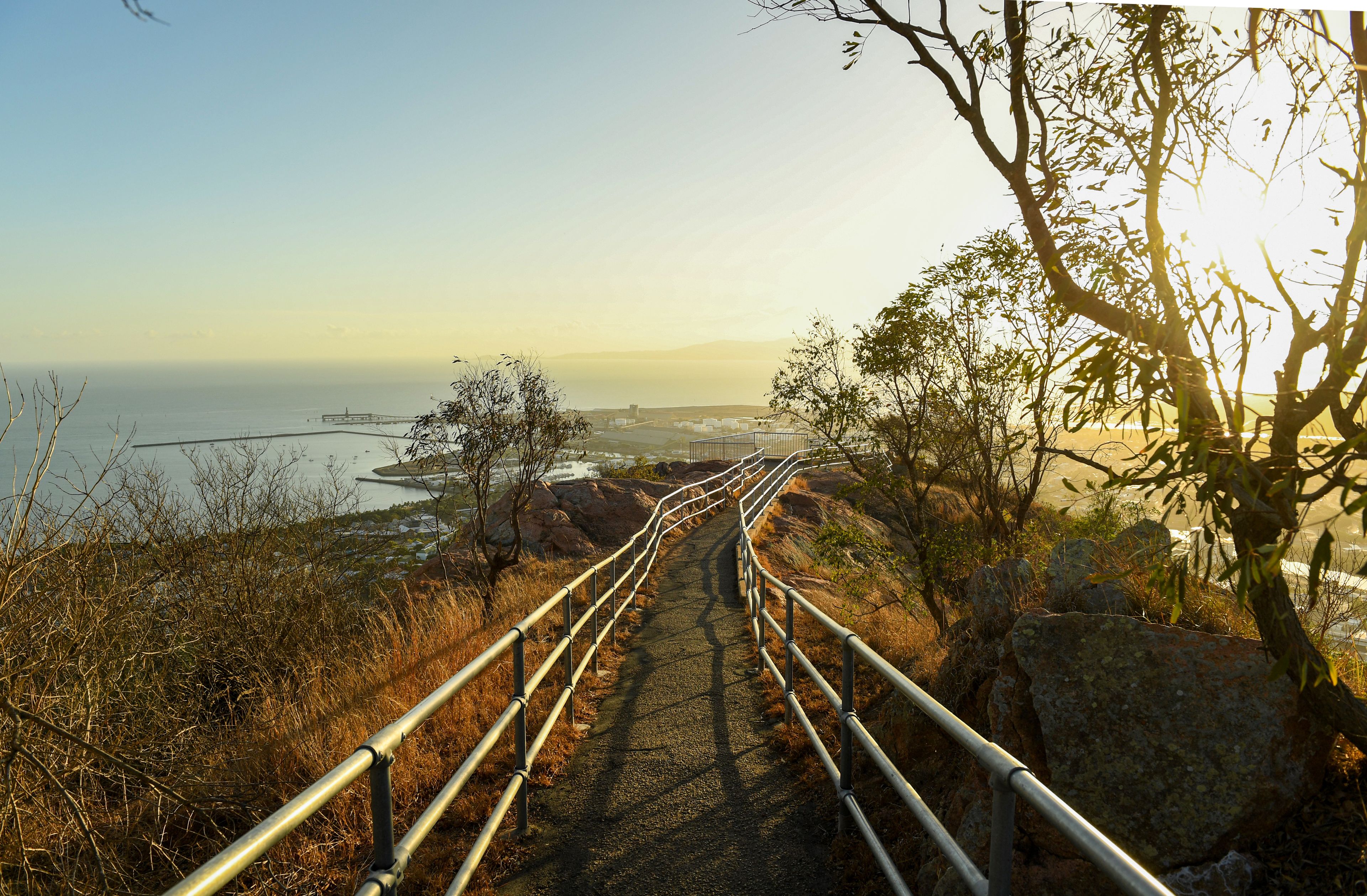 A coastal pathway with metal railings, leading towards a distant shoreline with a pier and buildings.