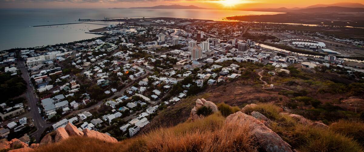 A coastal cityscape at sunset with a prominent bridge, dense residential area, and a hilly terrain in the foreground.