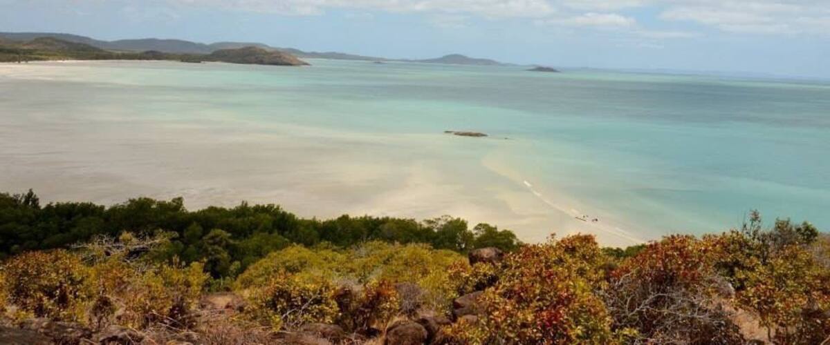 A coastal landscape with a sandy beach, clear turquoise waters, and distant hills under a partly cloudy sky.