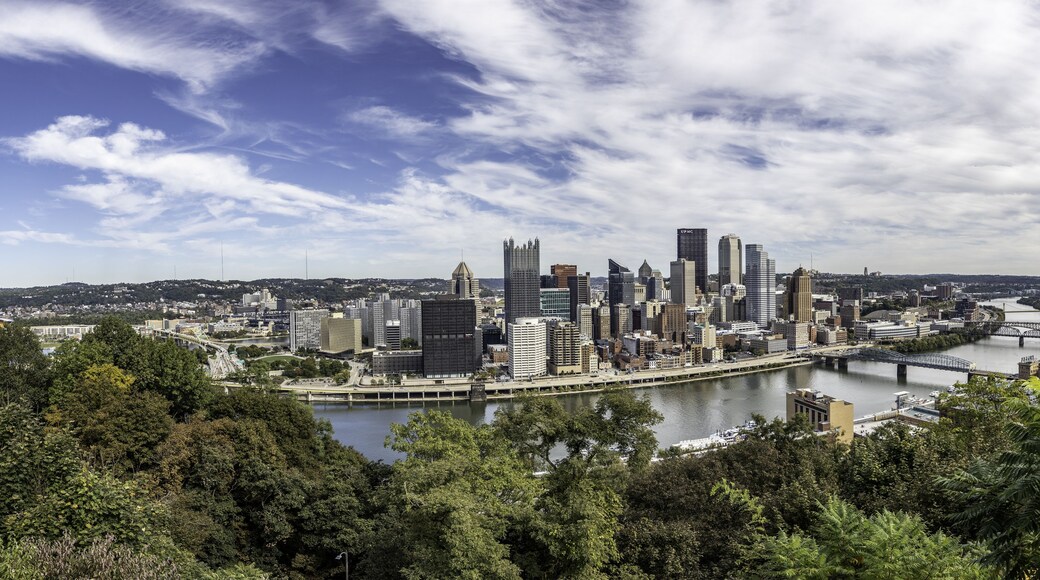 Aerial view of the Pittsburgh skyline with skyscrapers, river confluence, bridges, and lush green foliage in the foreground under a bright sky in Pittsburgh, Pennsylvania, United States.