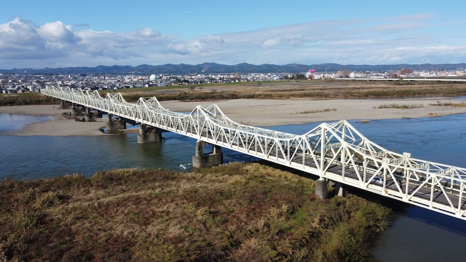 A white steel bridge over a river with a cityscape in the background.