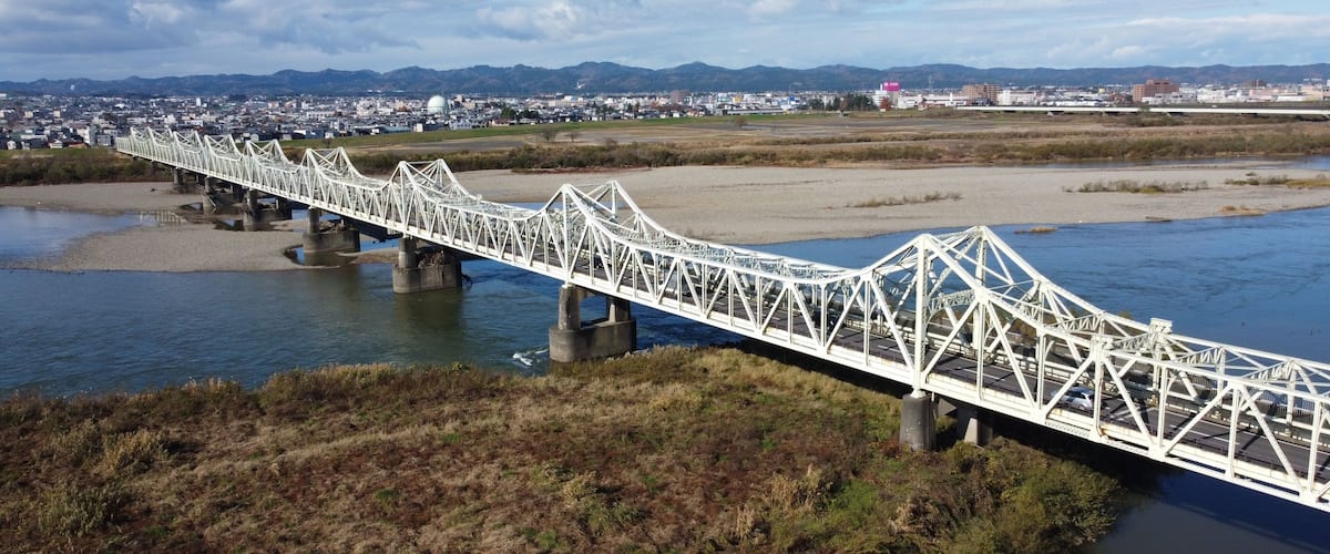A white steel bridge over a river with a cityscape in the background.