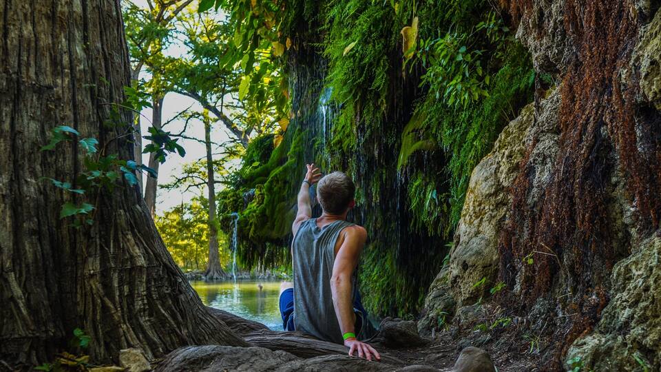 A person sitting on a log by a waterfall surrounded by trees and moss.