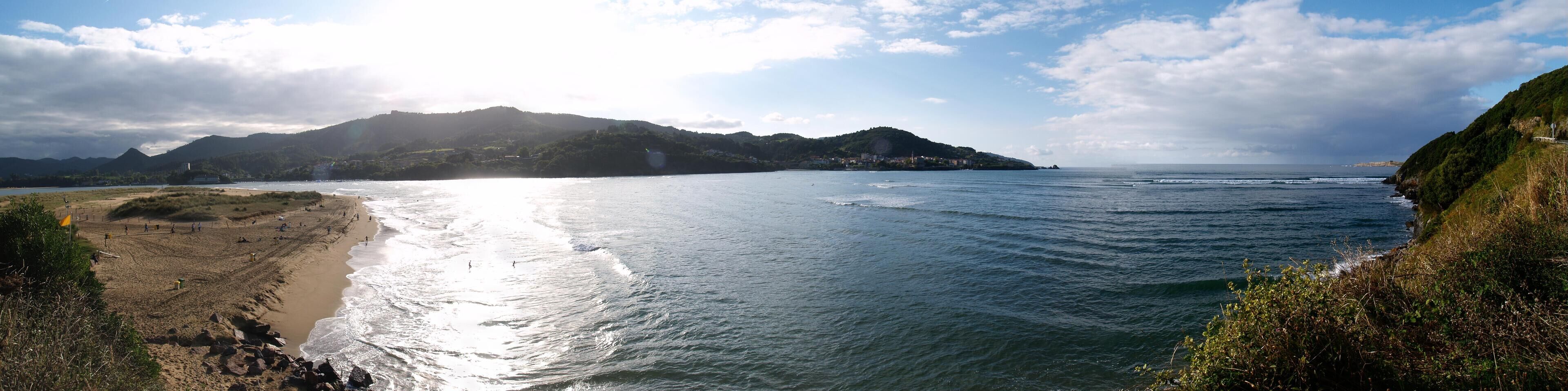 A coastal landscape with a sandy beach, calm sea, and a hill with vegetation.