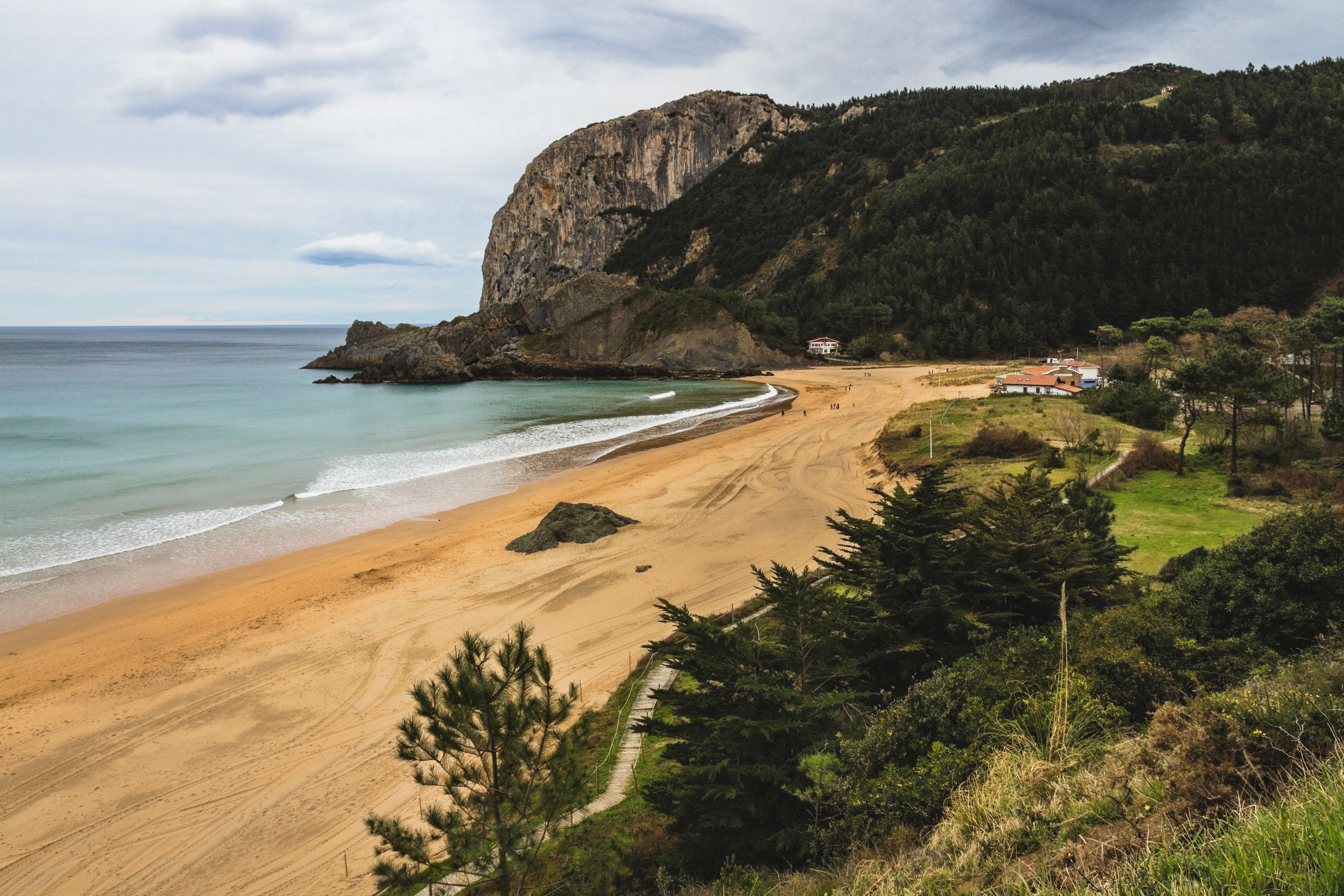 A coastal landscape with a sandy beach, a rocky cliff, and a few buildings in the distance.
