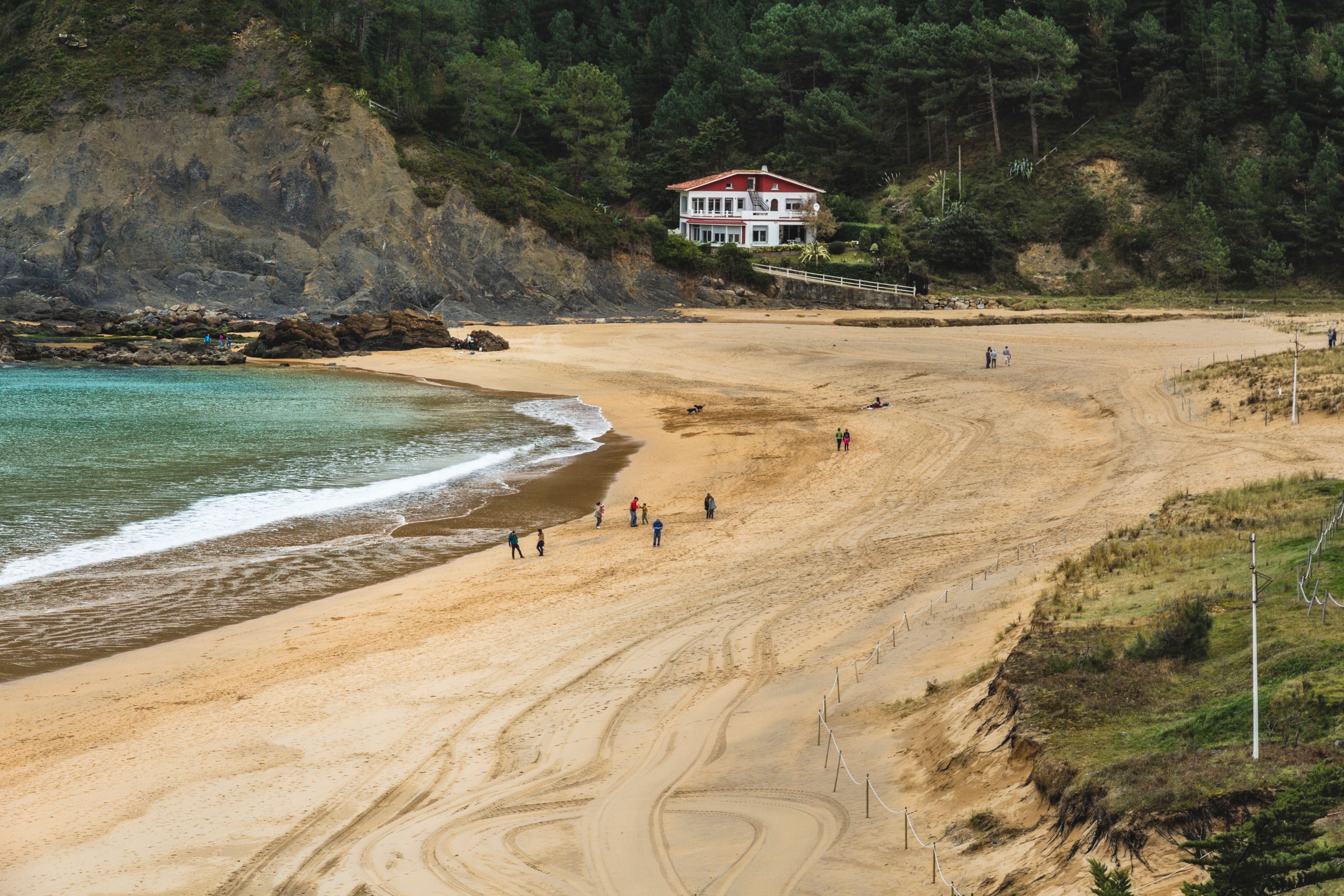 A sandy beach with footprints, a rocky cliff, and a red-roofed building.