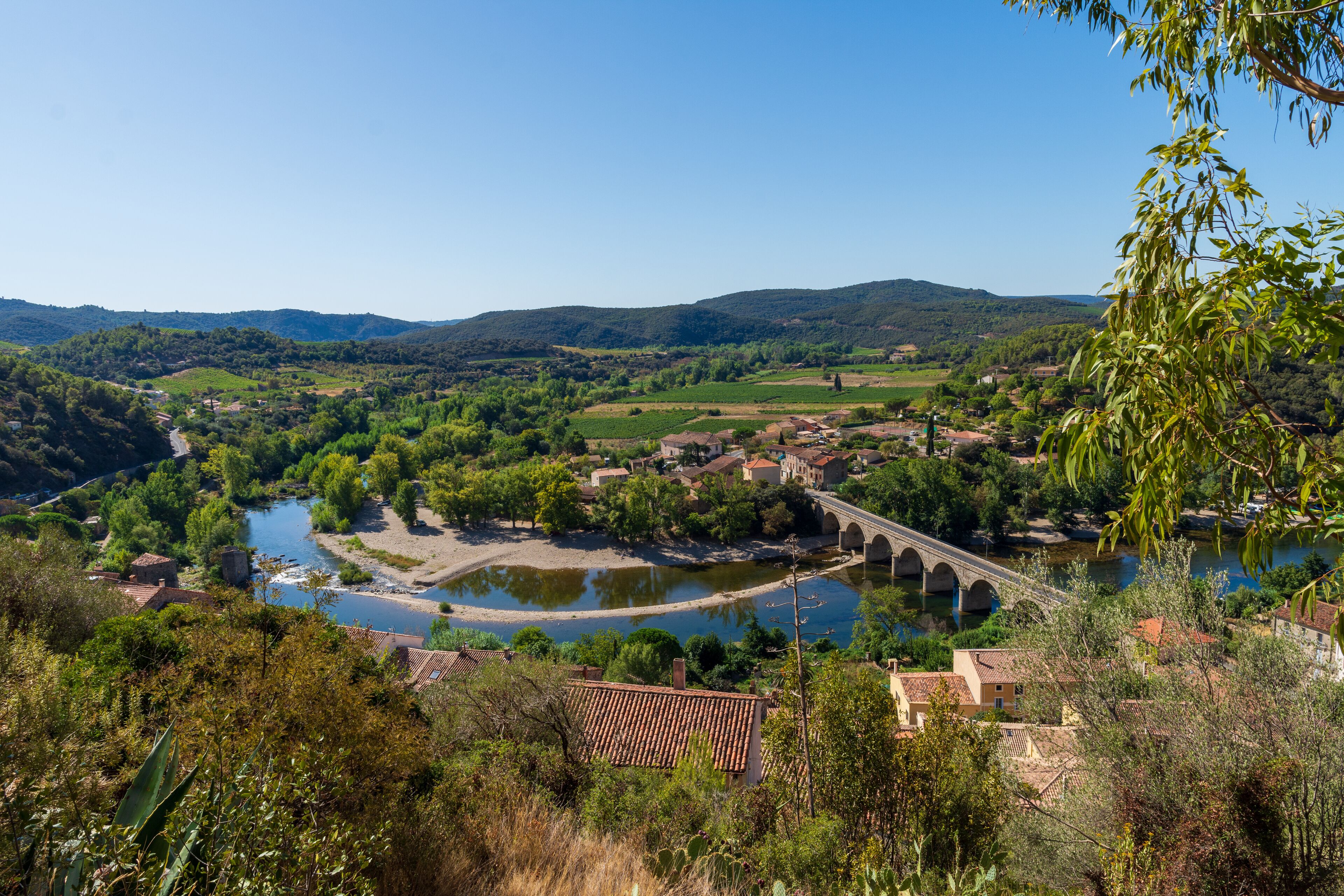 A river flowing through a valley with a bridge, surrounded by greenery and buildings.