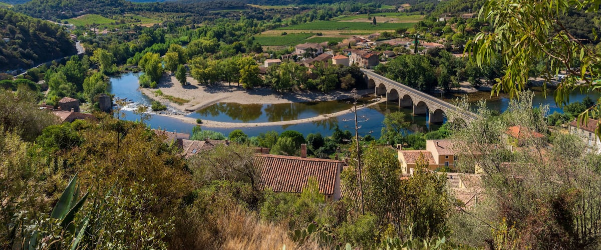 A river flowing through a valley with a bridge, surrounded by greenery and buildings.