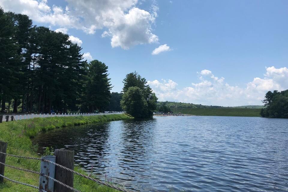 A calm lake with a wooden fence and trees on the left, and a clear sky with clouds.