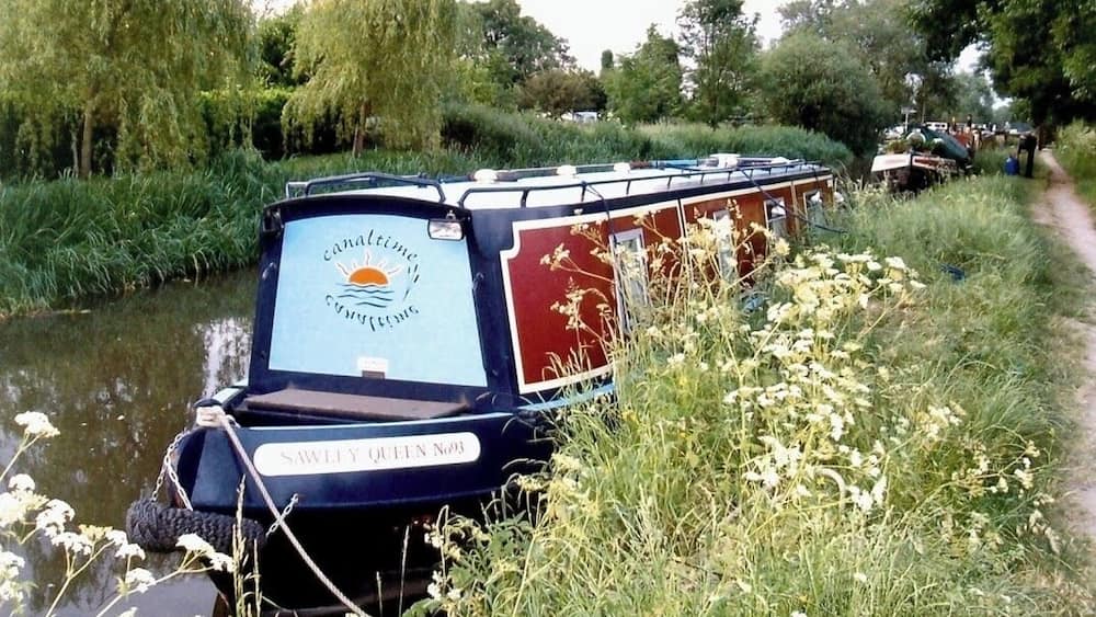 Une péniche nommée « Sawly Queen No. 3 » était amarrée au bord d’une rivière, avec une végétation luxuriante et un sentier visible au loin.