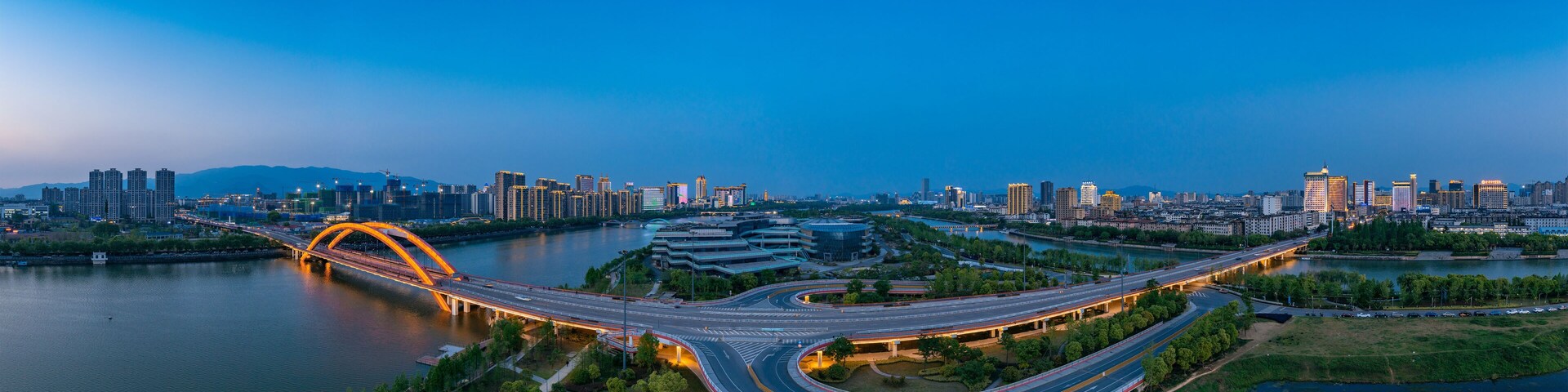 Un paisaje urbano con un puente curvo destacado sobre un río, rodeado de edificios altos y una autopista bien iluminada.