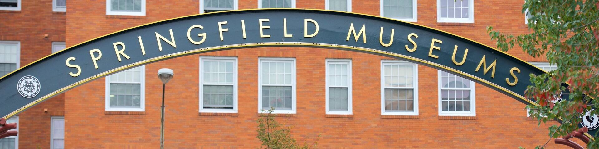A sign for Springfield Museums arching over a brick building with multiple windows.