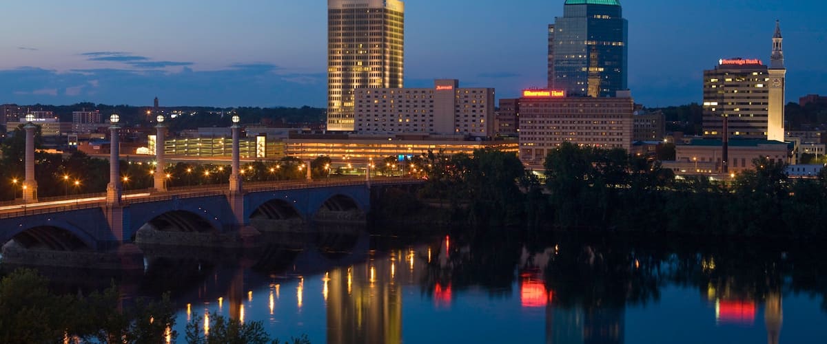 A city skyline at dusk with illuminated buildings, a bridge, and a river reflecting lights.