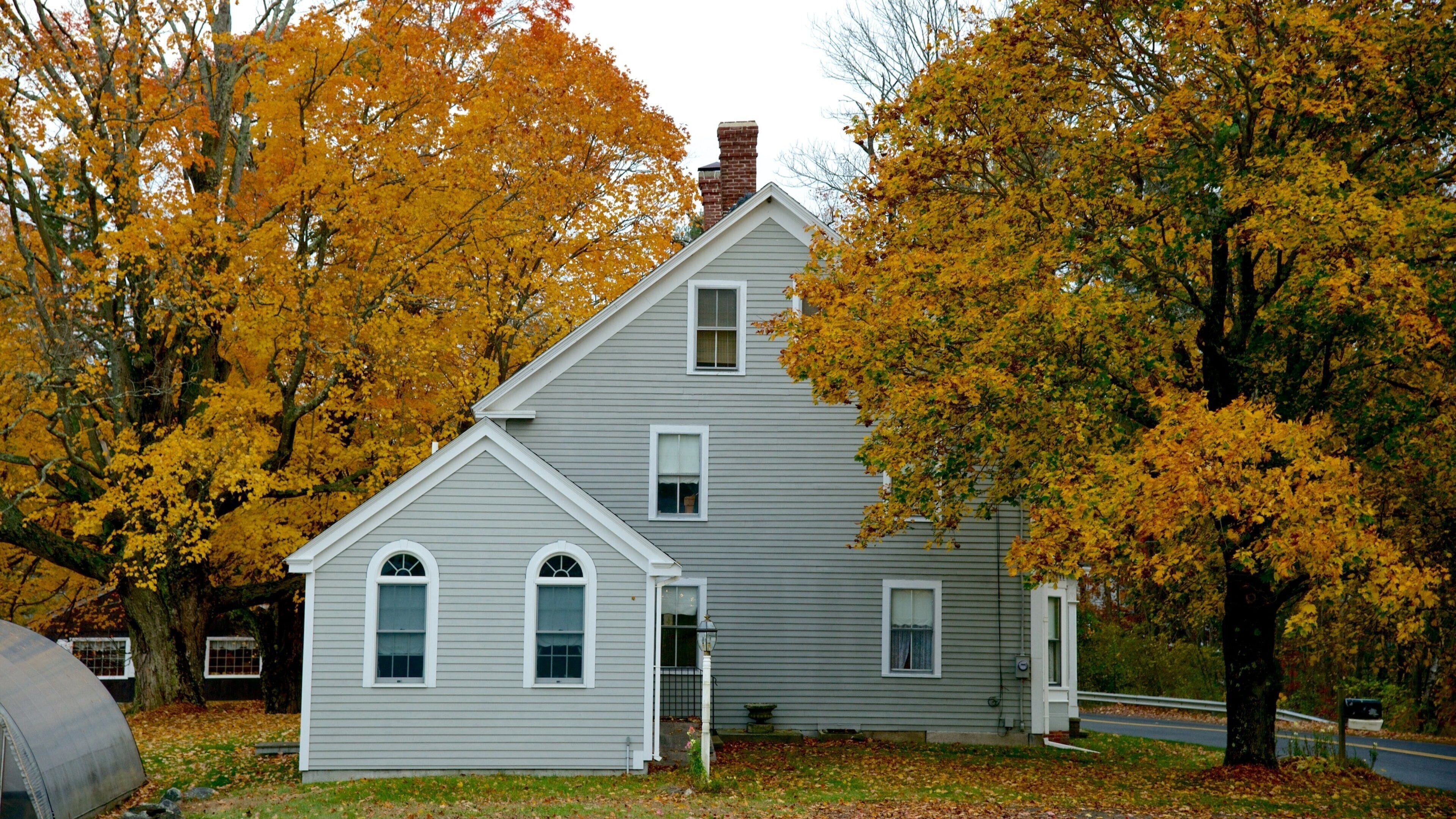 A two-story house with a gray exterior and white trim, surrounded by trees with yellow autumn leaves.