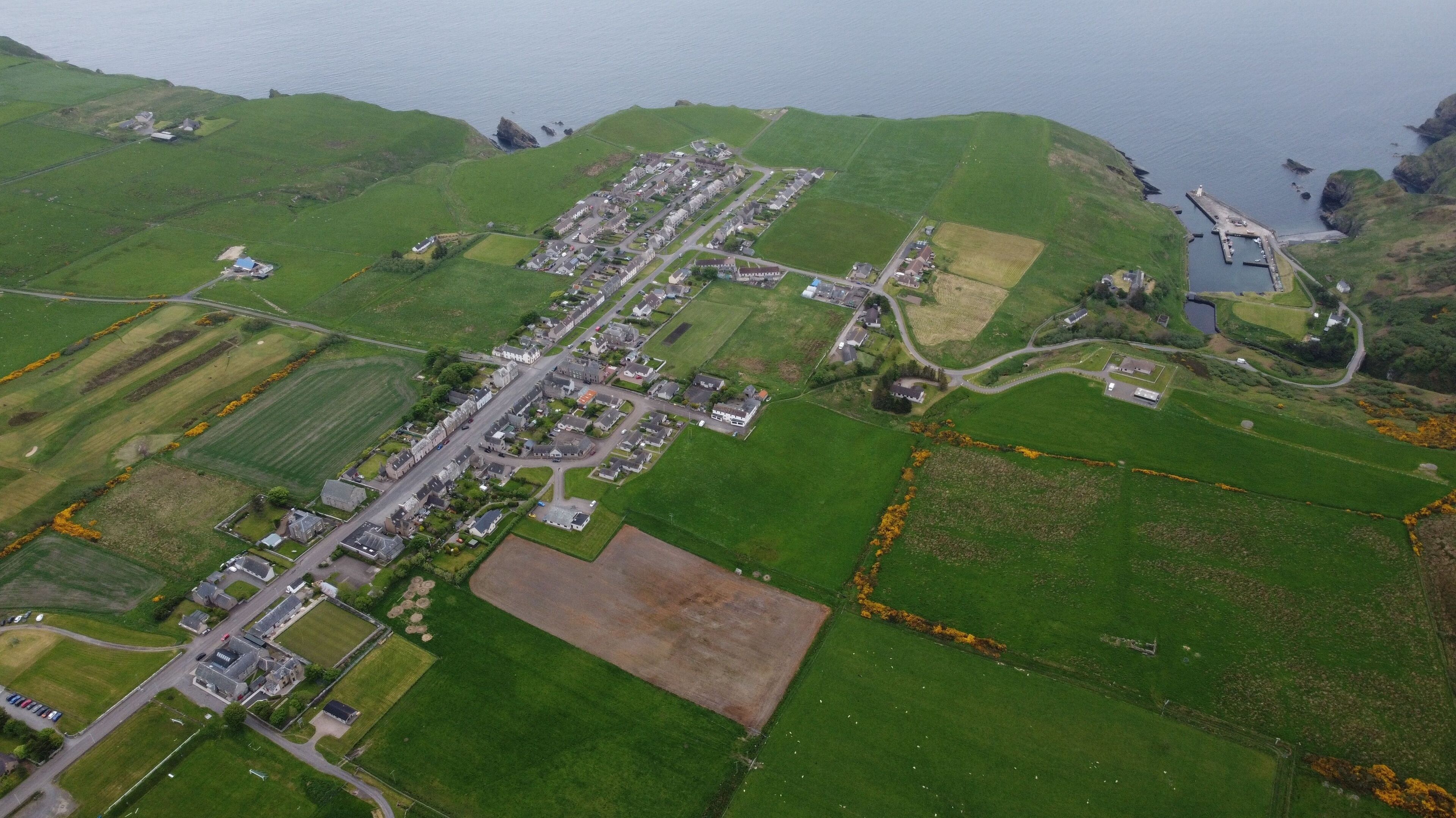 An aerial view of a coastal village with green fields, a main road, and a small harbor.