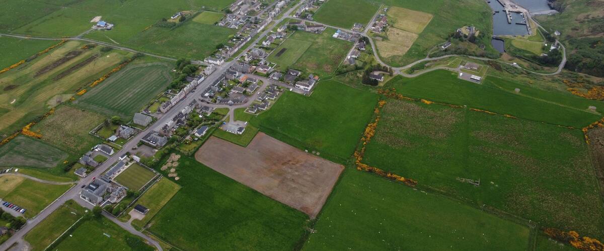 An aerial view of a coastal village with green fields, a main road, and a small harbor.