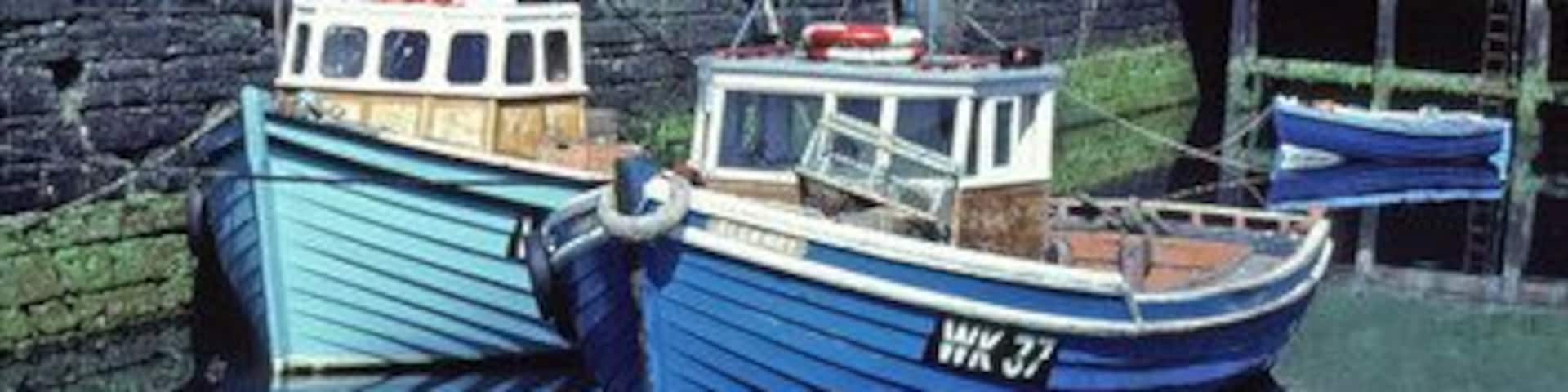 Two boats docked at a harbor with a stone wall and a grassy hill in the background.