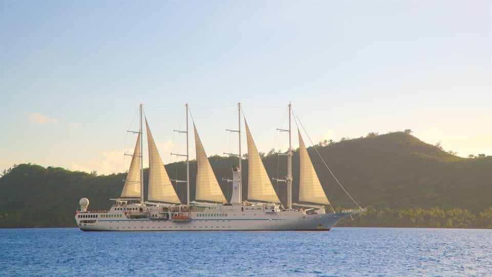 A large sailing ship with multiple sails on a calm body of water, with a hilly landscape in the background.
