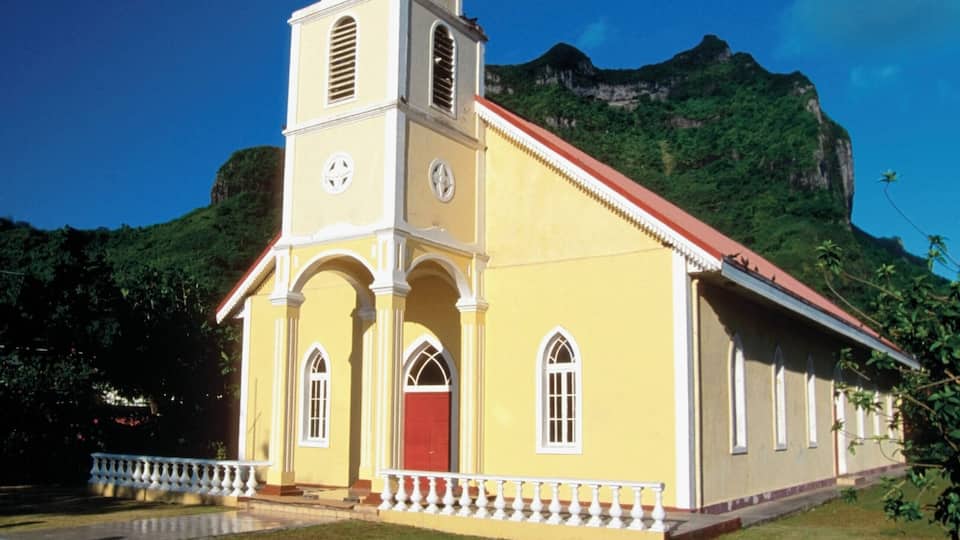 A yellow church with a red steeple and a clock tower, surrounded by lush greenery and mountains.