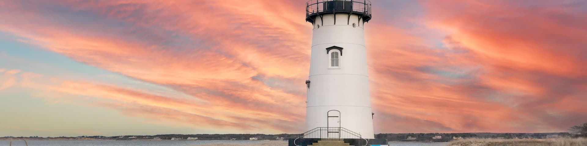 A white lighthouse with a black top, situated on a sandy beach with a backdrop of a vibrant sunset sky.