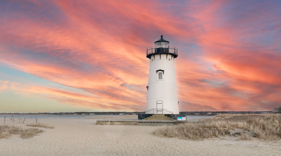 Un faro bianco con la sommità nera, situato su una spiaggia sabbiosa con un cielo al tramonto dai colori vivaci.