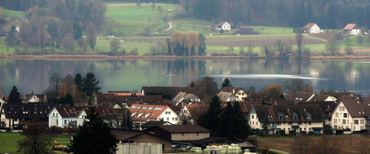 A lakeside village with houses, a calm lake, and rolling hills in the background.
