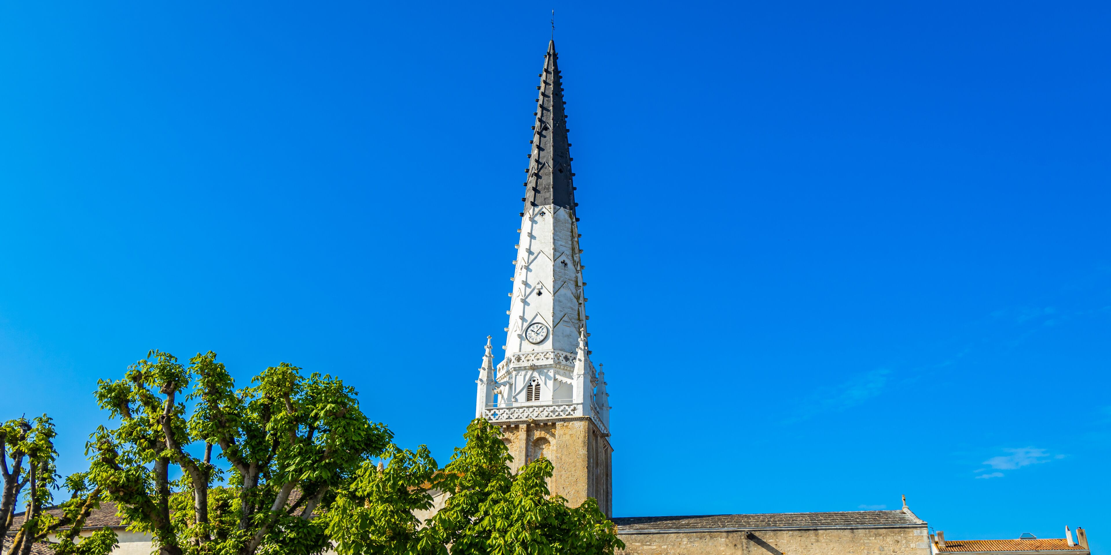 A tall church tower with intricate designs and a clock, surrounded by trees under a clear blue sky.