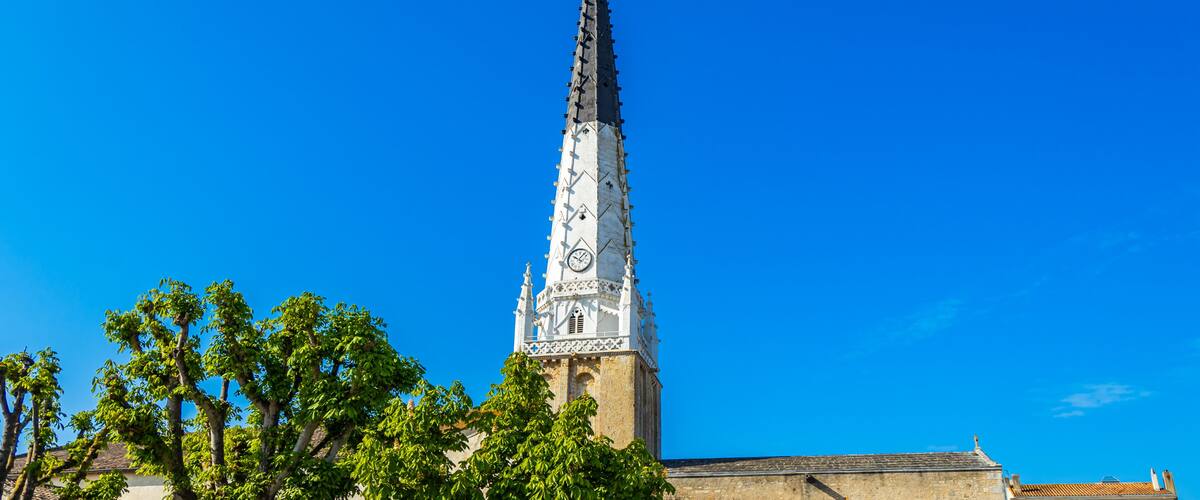 Un'alta torre campanaria con decorazioni elaborate e un orologio, circondata da alberi sotto un cielo azzurro limpido.