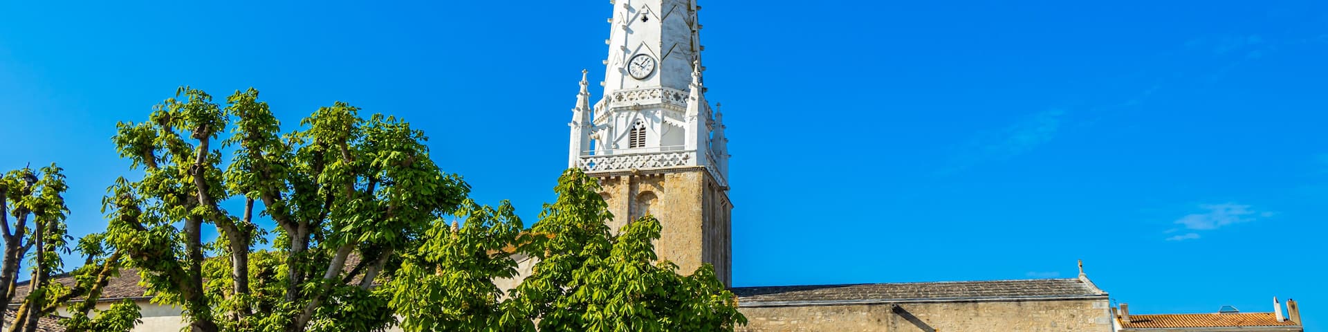A tall church tower with intricate designs and a clock, surrounded by trees under a clear blue sky.