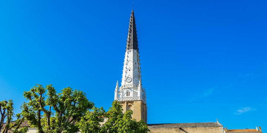 Un'alta torre campanaria con decorazioni elaborate e un orologio, circondata da alberi sotto un cielo azzurro limpido.