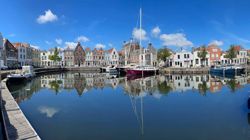 A canal with boats, a tall sailing ship, and historic buildings reflecting on the water.