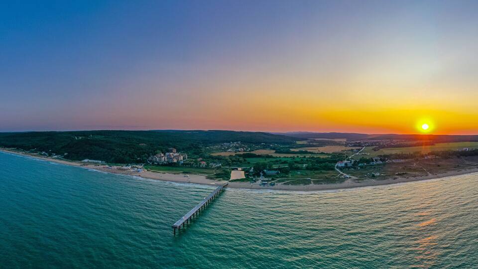 A coastal sunset with a pier extending into the sea, surrounded by hills and a small settlement.