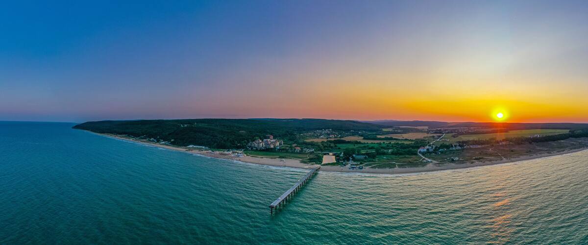 A coastal sunset with a pier extending into the sea, surrounded by hills and a small settlement.