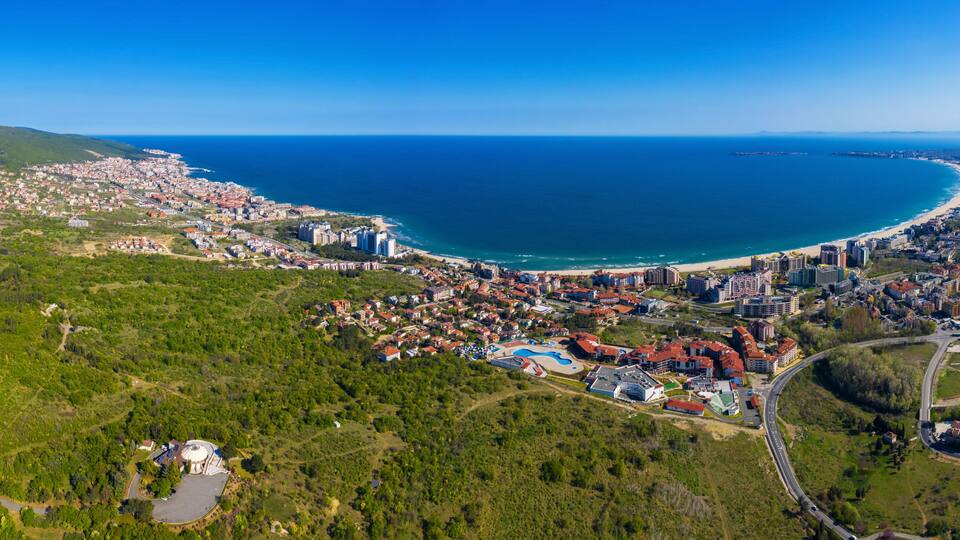An aerial view of a coastal city with a beach, dense urban area, and surrounding greenery.