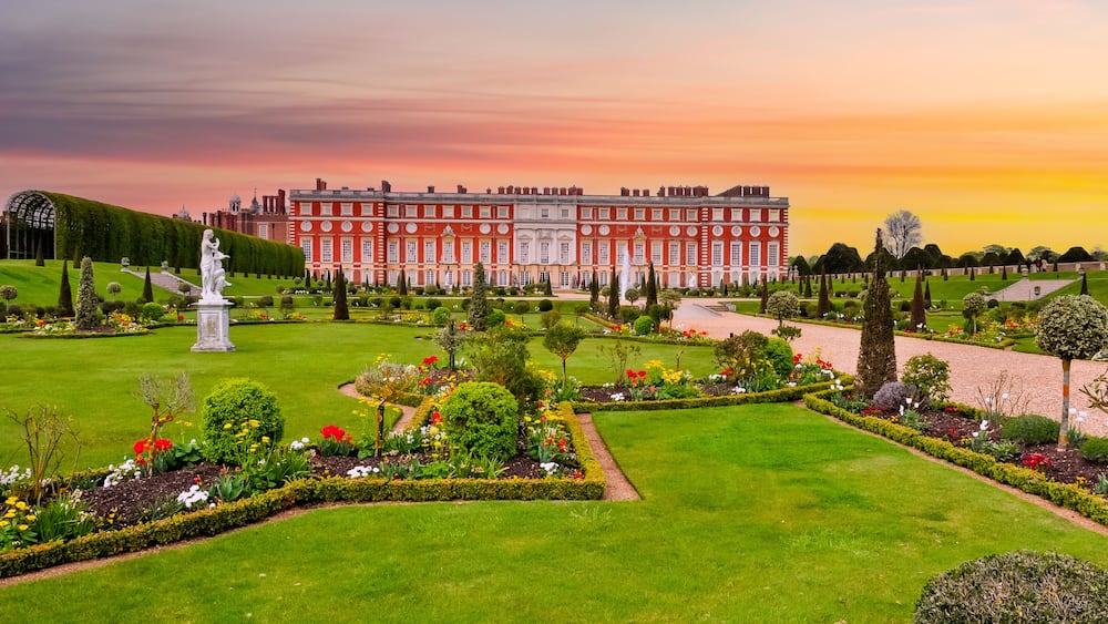 A grand palace with a well-manicured garden, featuring a statue, neatly trimmed hedges, and a variety of colorful flowers.