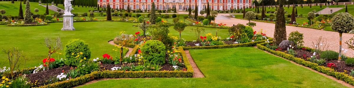 A grand palace with a well-manicured garden, featuring a statue, neatly trimmed hedges, and a variety of colorful flowers.