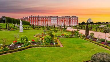 A grand palace with a well-manicured garden, featuring a statue, neatly trimmed hedges, and a variety of colorful flowers.
