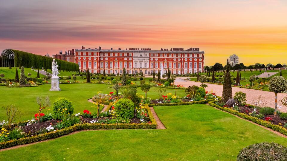 A grand palace with a well-manicured garden, featuring a statue, neatly trimmed hedges, and a variety of colorful flowers.