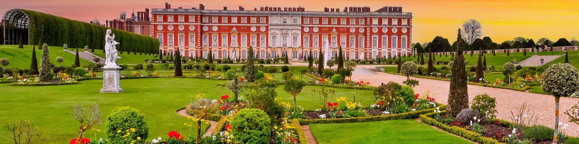 Um palácio imponente com um jardim bem cuidado, apresentando uma estátua, sebes aparadas e uma variedade de flores coloridas.