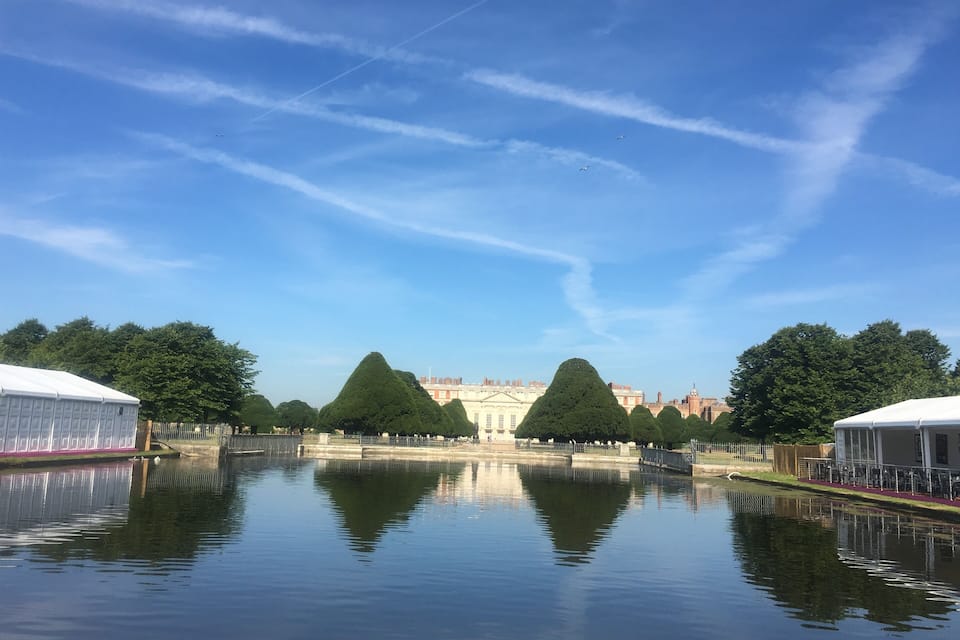 A calm water body reflecting trees and buildings, with a clear sky and contrails.
