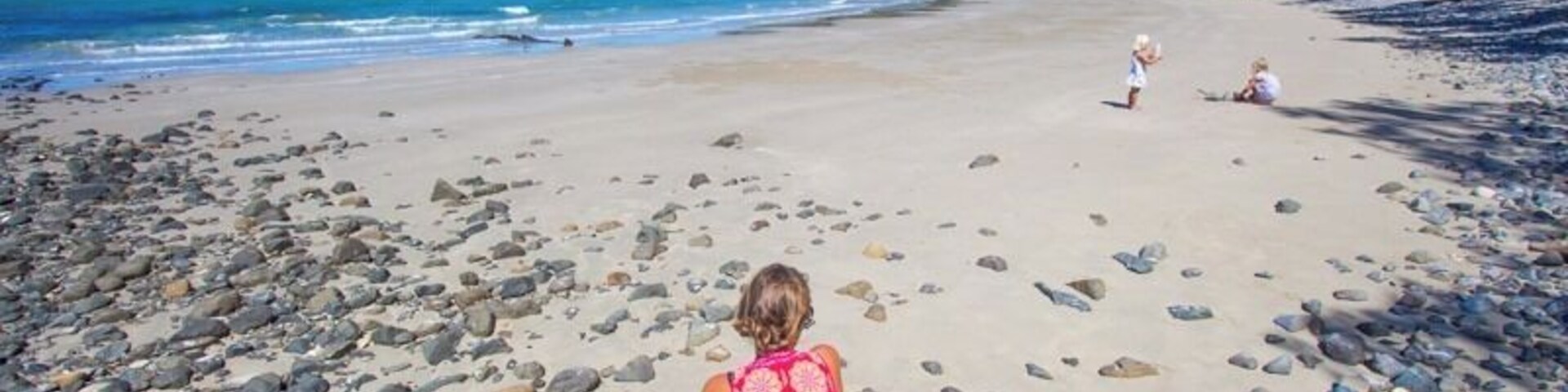 Mulher de vestido vermelho sentada numa praia rochosa, com céu azul límpido e mar calmo.