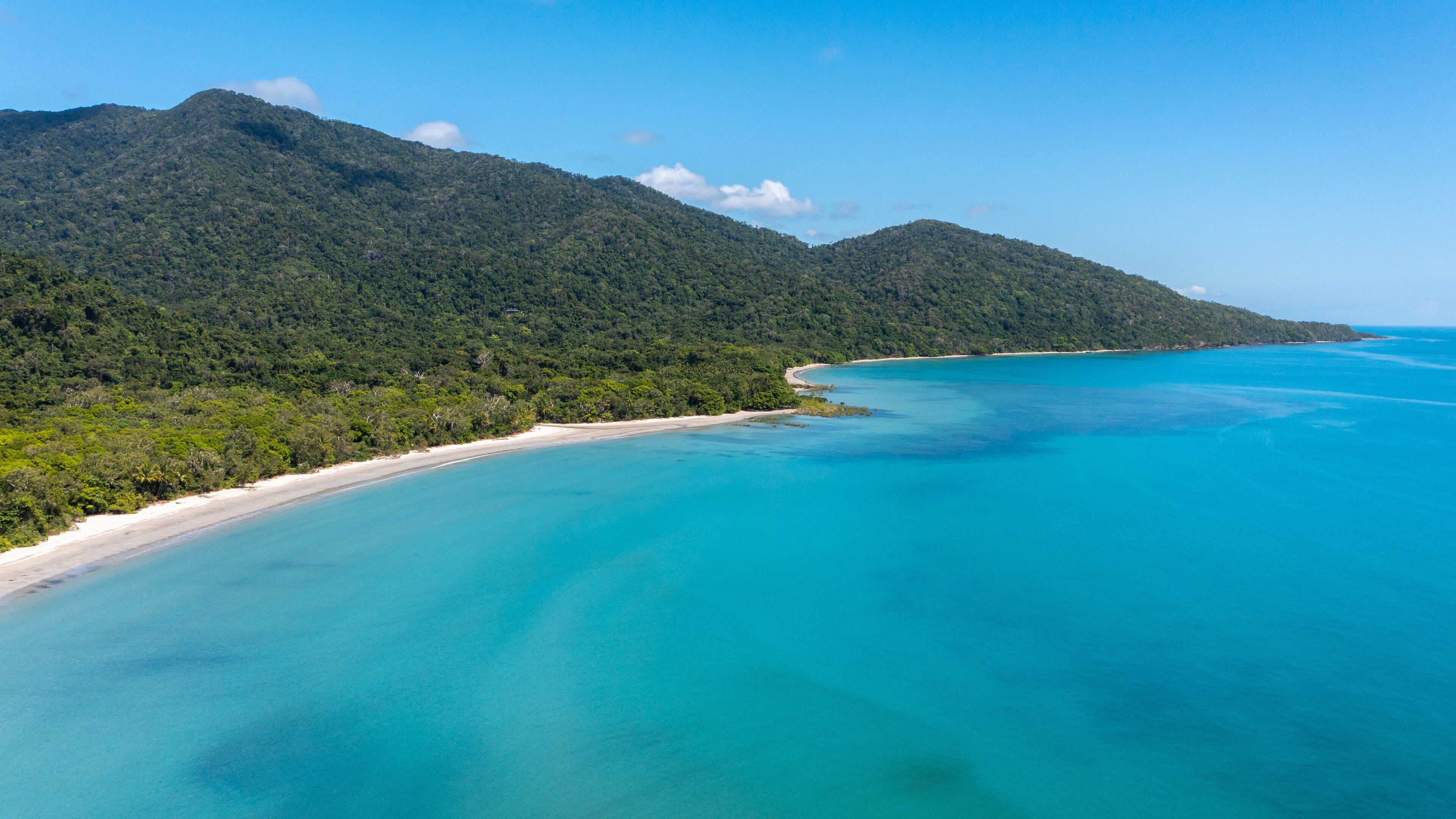 A coastal landscape with a beach, turquoise waters, and a forested mountain.