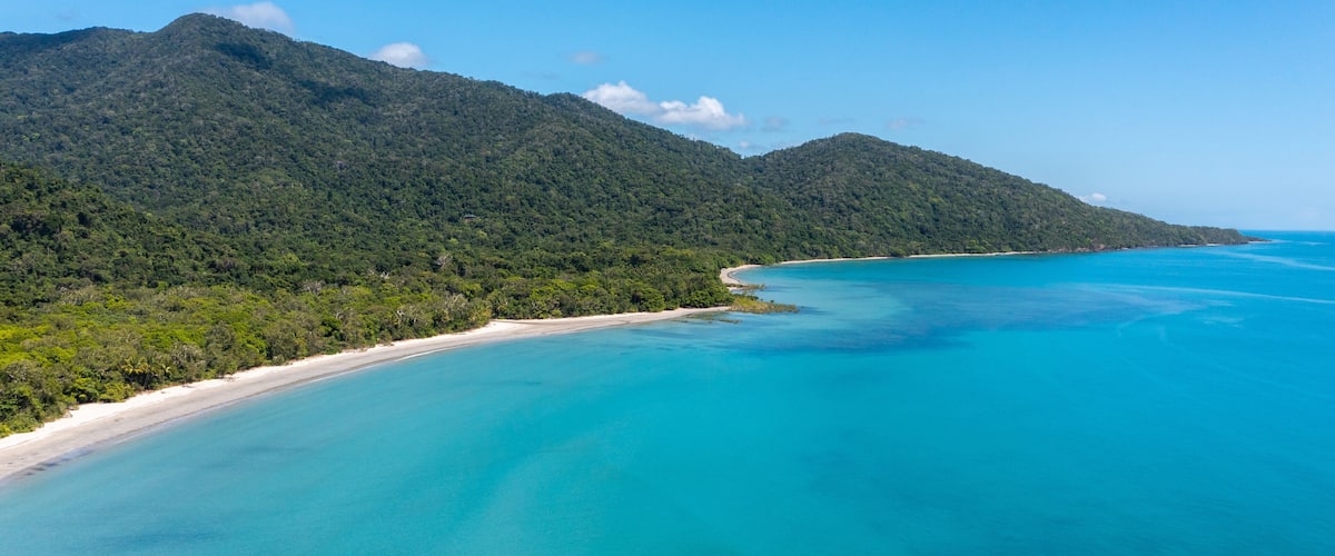 A coastal landscape with a beach, turquoise waters, and a forested mountain.