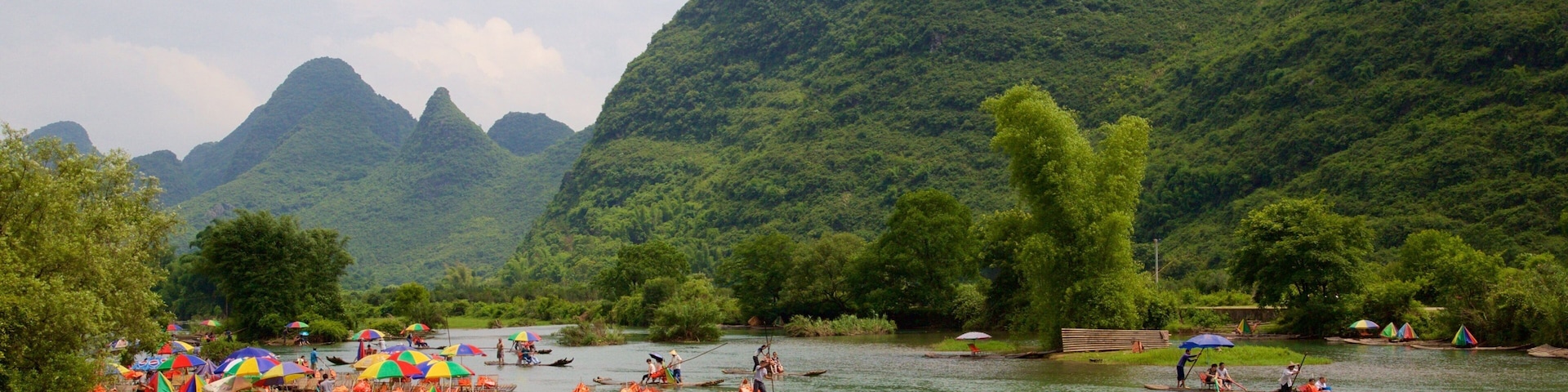 A river with boats and colorful umbrellas, surrounded by lush green mountains.