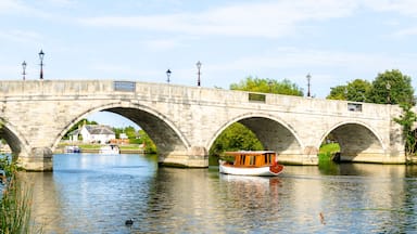 Uma ponte de pedra com arcos sobre um rio calmo, um barco de madeira e patos nadando.